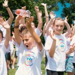 Group of cheerful children raising hands in a park during a sunny day, wearing t-shirts with event logos.